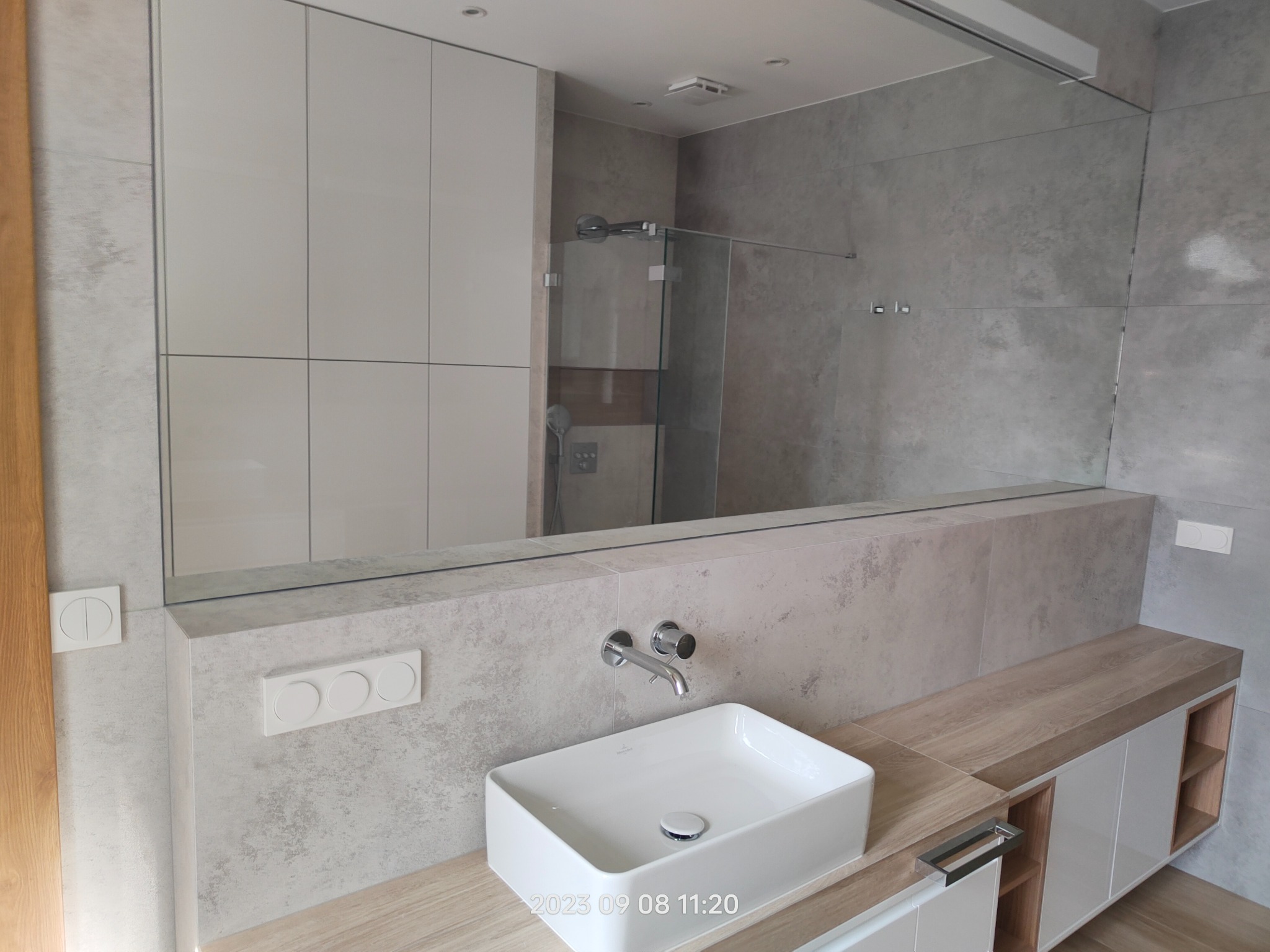 Nowoczesna łazienka w Warszawie z prostokątną umywalką Villeroy & Boch, zamontowaną na drewnianym blacie z szafkami, szare płytki na ścianach i duże lustro.