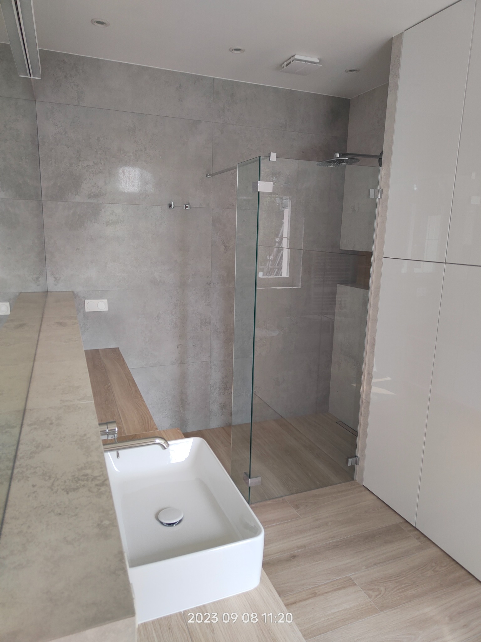 Nowoczesna łazienka z kabiną prysznicową typu walk-in, umywalką nablatową i szarą okładziną ścienną imitującą beton.