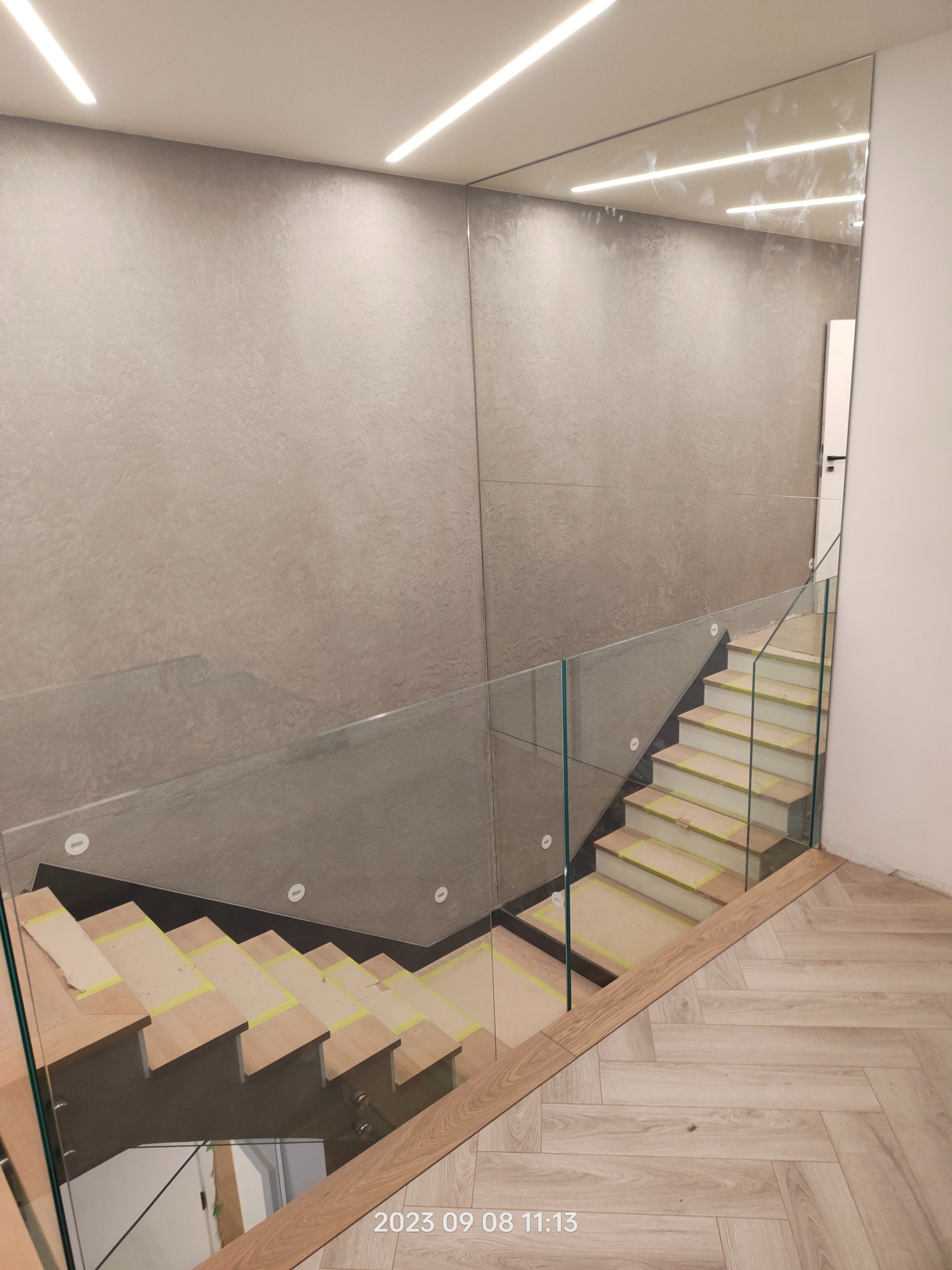 Nowoczesne schody drewniane z szklaną balustradą, zabezpieczone taśmą malarską podczas prac wykończeniowych, parkiet ułożony w jodełkę, ściana pokryta dekoracyjną fakturą.