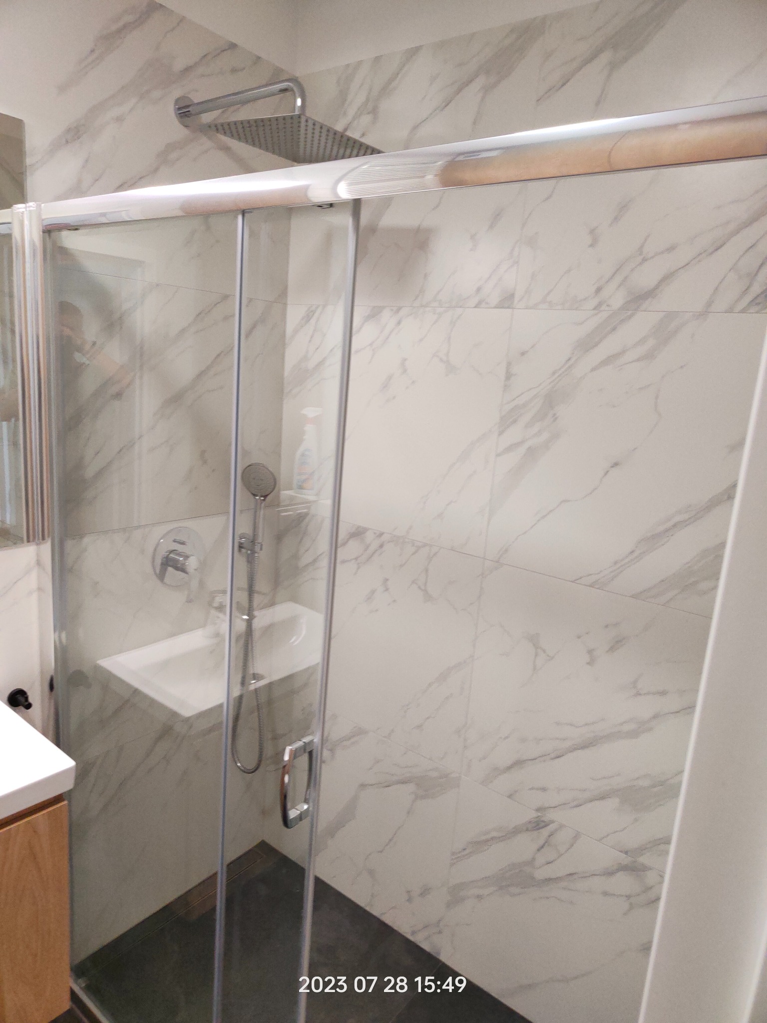 Nowoczesna kabina prysznicowa z przesuwnymi drzwiami, marmurowe płytki na ścianach, bateria podtynkowa i deszczownica. Widoczna umywalka i fragment szafki pod umywalką.