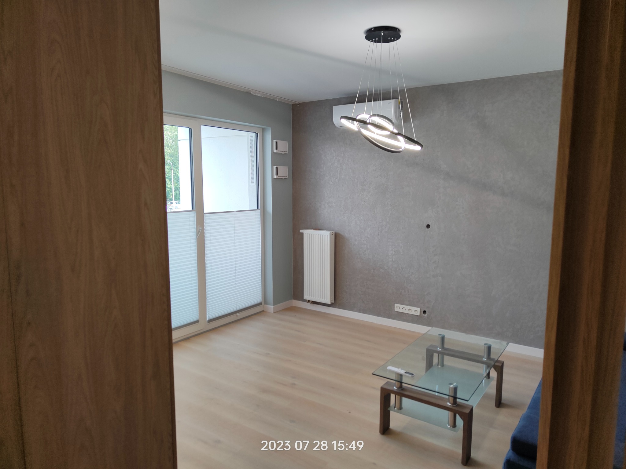 Nowoczesny salon z szarą ścianą o fakturze, designerską lampą wiszącą i szklanym stolikiem kawowym, widok zza drewnianej framugi drzwi.