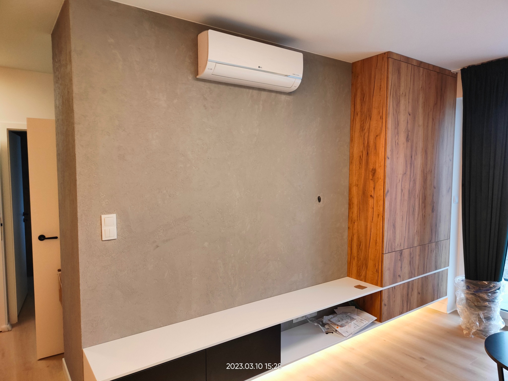 Szara ściana z dekoracyjną strukturą, zamontowany klimatyzator LG, wbudowana szafka z drewnopodobnym fornirem, biało-czarna szafka RTV z podświetleniem LED na podłodze z paneli.