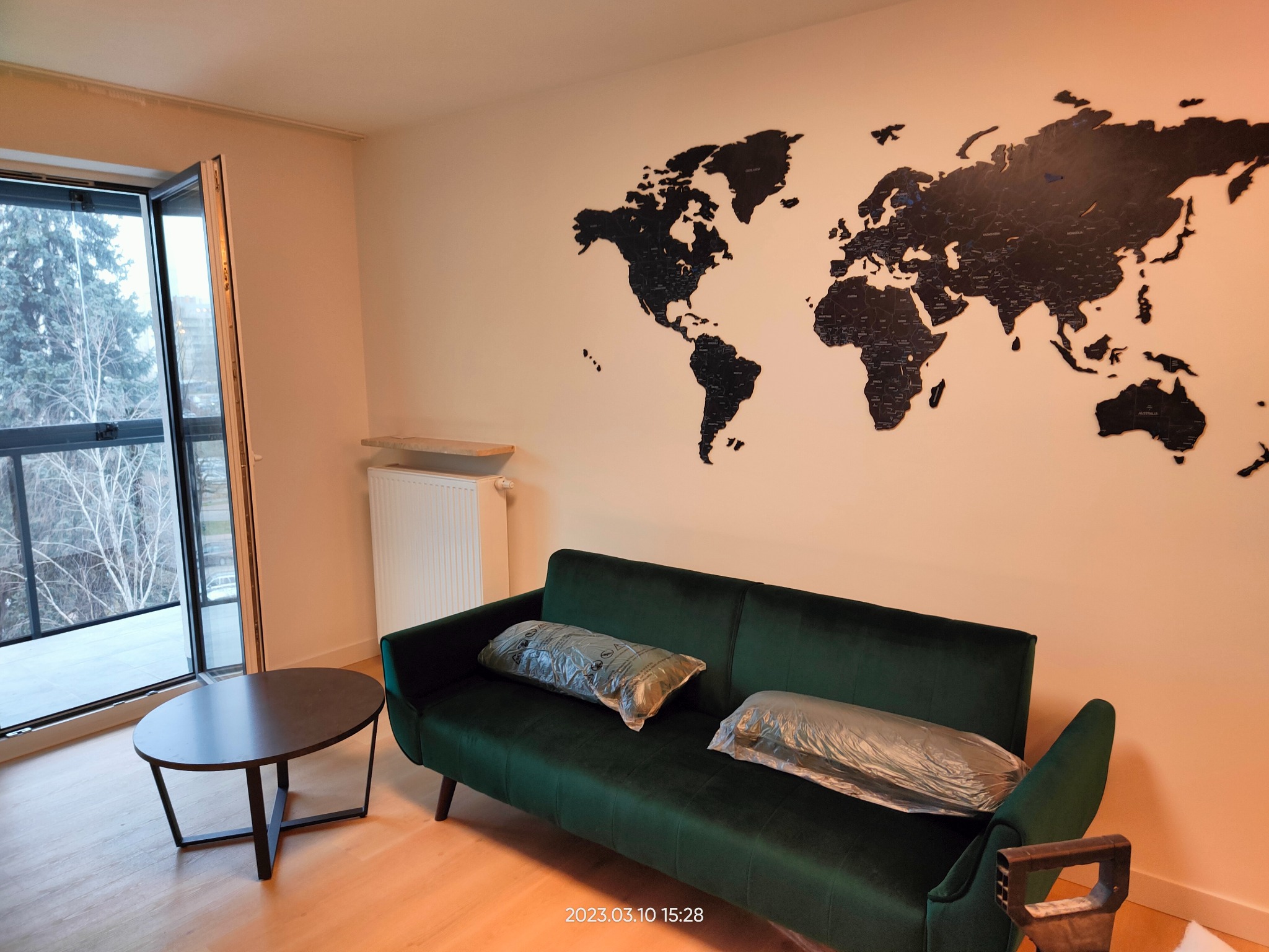 Wnętrze pokoju po remoncie: zielona sofa z poduszkami w folii, stolik kawowy, mapa świata na ścianie, otwarte drzwi balkonowe z widokiem na drzewa.