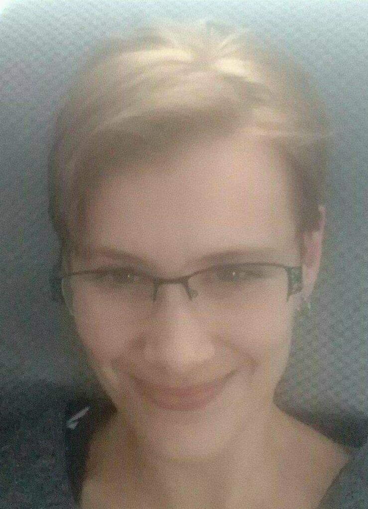 Portret uśmiechniętej kobiety z krótkimi blond włosami i okularami w cienkich oprawkach, sfotografowanej z bliska na rozmytym tle.