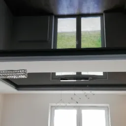 Czarny, błyszczący sufit napinany z oknem odbijającym się w jego powierzchni, widoczne nowoczesne oświetlenie i fragment ściany z oknem.