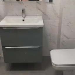 Nowoczesna łazienka z szarym zestawem umywalkowym z dwiema szufladami, białą toaletą i kabiną prysznicową z grafitowymi szybami, wykończona białymi płytkami imitującymi marmur.