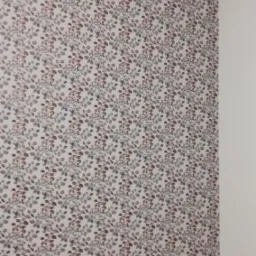 Ściana w pokoju pokryta tapetą w drobny wzór liści w odcieniach szarości, brązu i bieli. Widoczny fragment drewnianej podłogi i białe listwy przypodłogowe.