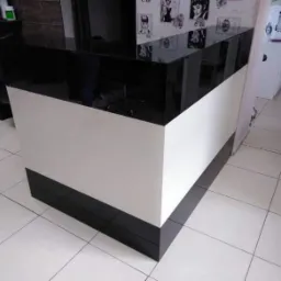 Czarno-biała lada recepcyjna z błyszczącym blatem, widoczna w salonie fryzjerskim z białymi płytkami na podłodze i tapetą z portretami na ścianie w tle.