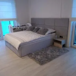 Szare łóżko tapicerowane z pikowanym zagłówkiem, obok szafka nocna, puszysty dywanik i łóżeczko dziecięce w tle, w jasnym pomieszczeniu z drewnianą podłogą.