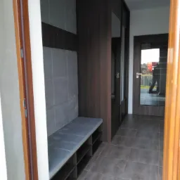 Wnętrze przedpokoju z zabudowaną szafą w ciemnym drewnie, tapicerowanym siedziskiem i panelem ściennym w szarym odcieniu oraz otwartymi półkami na buty.