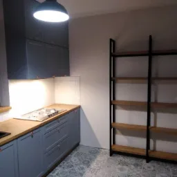 Niebieska zabudowa kuchenna z drewnianym blatem, miedzianymi uchwytami i płytą grzewczą, uzupełniona o regał z drewnianymi półkami na tle wzorzystej podłogi.