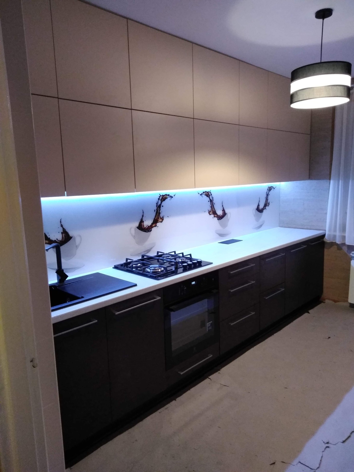 Nowoczesna kuchnia z zabudową w odcieniach beżu i brązu, z białym blatem i podświetlanym panelem szklanym z motywem kawy, oświetlona lampą wiszącą.