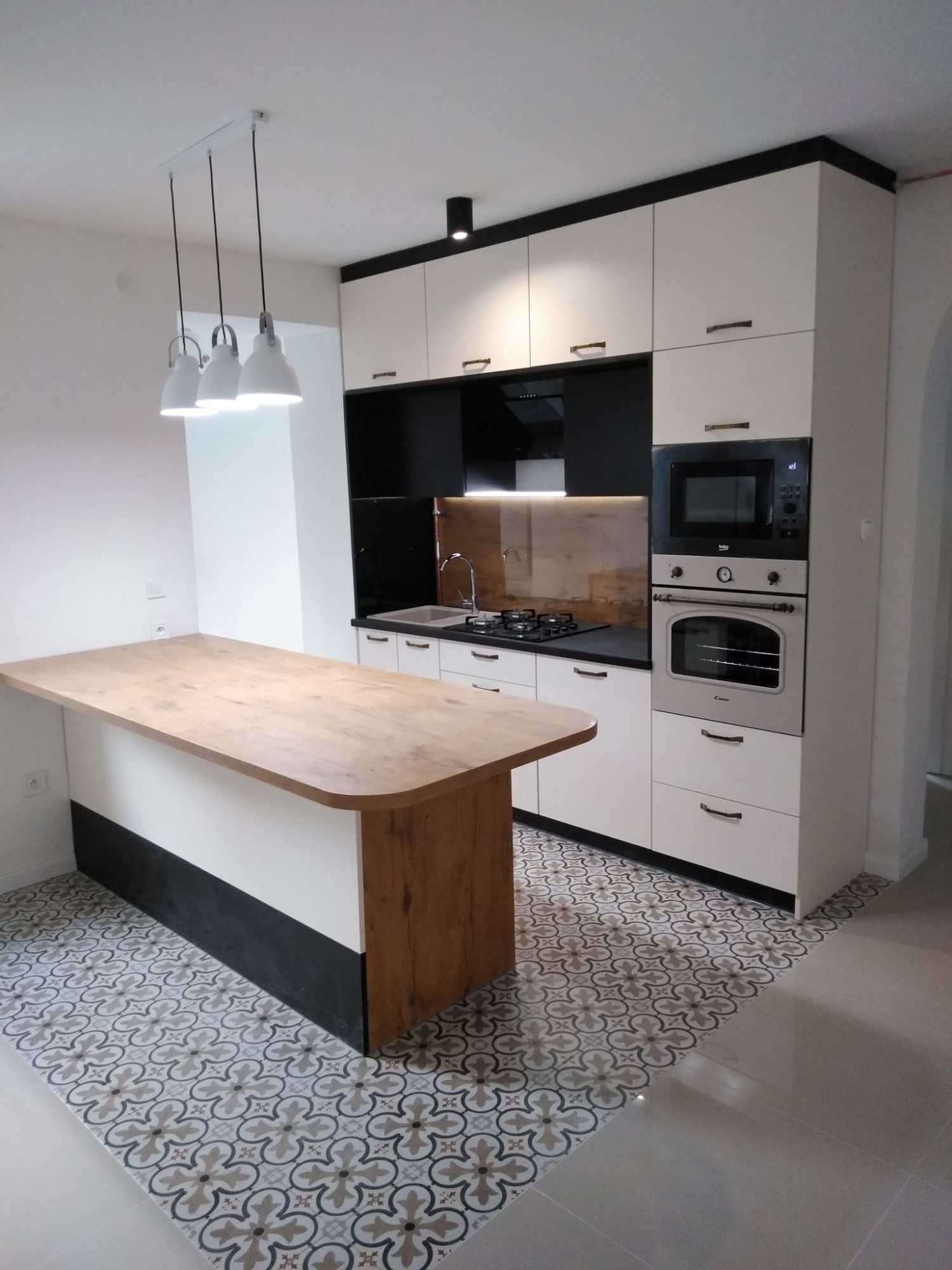 Nowoczesna kuchnia z wyspą, białe szafki z czarnymi akcentami, drewniany blat wyspy, wzorzysta podłoga, oświetlenie punktowe i wiszące lampy.