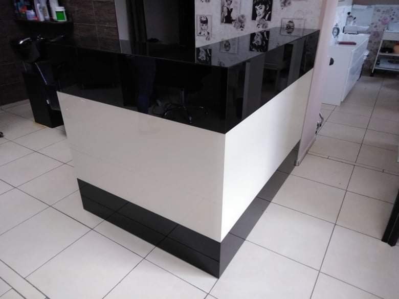 Czarno-biała lada recepcyjna z błyszczącym blatem, widoczna w salonie fryzjerskim z białymi płytkami na podłodze i tapetą z portretami na ścianie w tle.