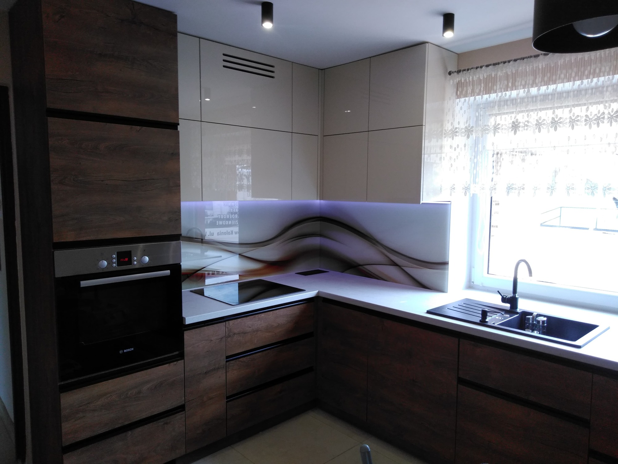 Nowoczesna kuchnia z frontami w odcieniach drewna i beżu, białym blatem oraz dekoracyjnym panelem nad blatem, oświetlona punktowymi lampami sufitowymi.