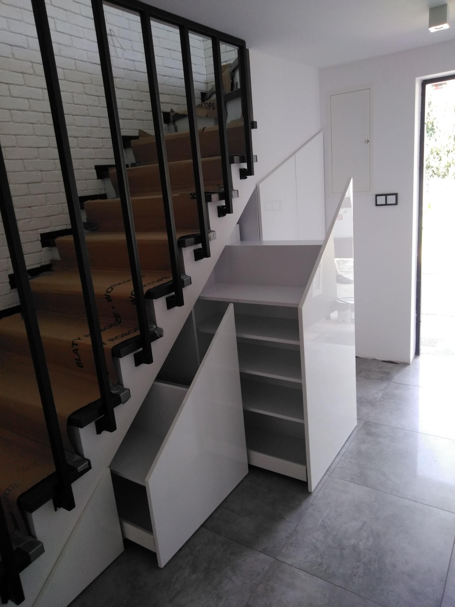 Białe szafki wnękowe pod schodami z czarną balustradą, widok z dołu na szarych płytkach podłogowych.