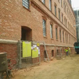 Renowacja ceglanej elewacji budynku z użyciem rusztowań i piaskarki, widoczny fragment ciężarówki i pracownik w żółtej kamizelce.