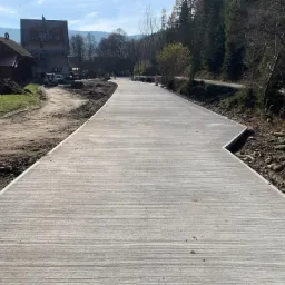 Nowo wybudowana, gładka, betonowa droga w górskim otoczeniu z widocznymi drzewami i budynkami w tle.