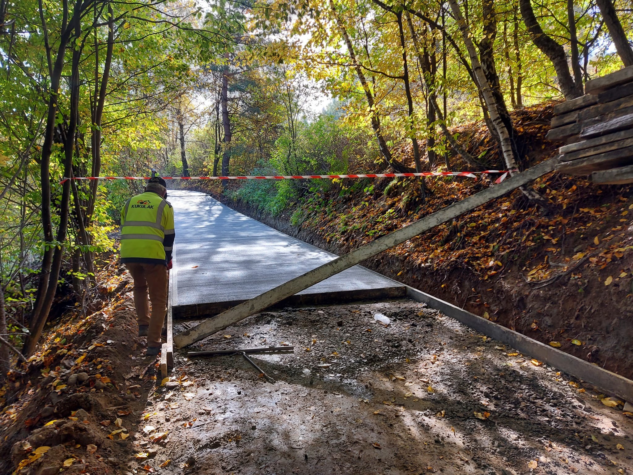 Świeżo wylana, betonowa droga leśna w trakcie realizacji, zabezpieczona czerwoną taśmą, widoczny pracownik w kamizelce odblaskowej i drewniane szalunki, otoczona jesiennym lasem.