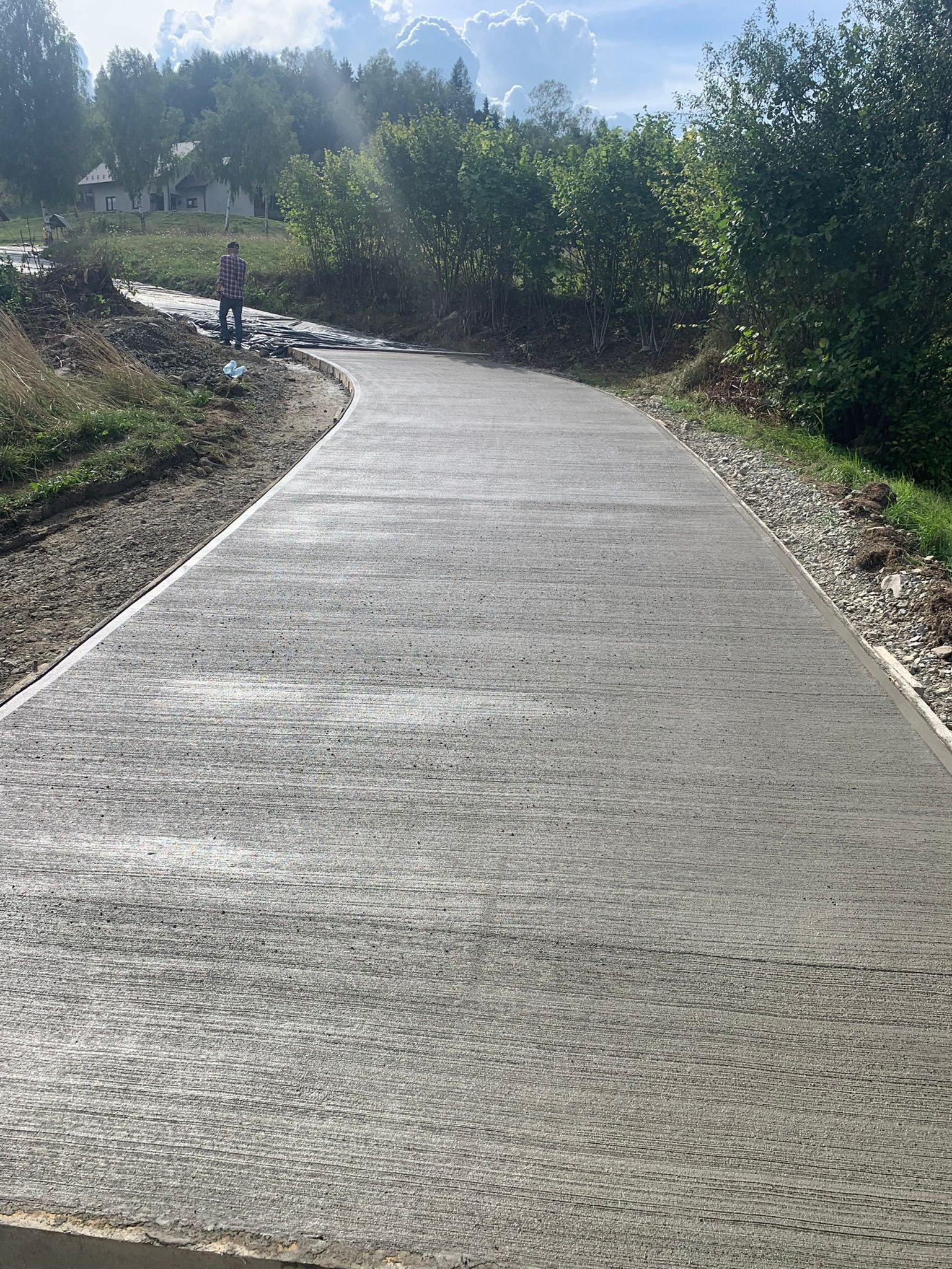 Nowo wylana, gładka, betonowa droga wijąca się przez wiejski krajobraz z osobą w oddali i świeżo posadzoną zielenią po bokach.