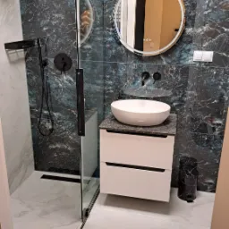 Nowoczesna łazienka z kabiną typu walk-in, umywalką na szafce z szufladami i okrągłym lustrem z podświetleniem. Płytki imitujące marmur w odcieniach granatu i bieli.
