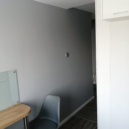 Wojewoda Jakub - Wnętrze pokoju po malowaniu ścian na szaro, widoczny stół z blatem drewnopodobnym i krzesło z metalowymi nogami, minimalistyczne oświetlenie sufitowe i biała zabudowa meblowa.