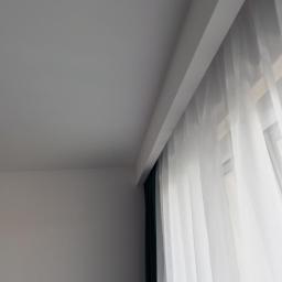 Wojewoda Jakub - Fragment pokoju po remoncie: gładka, pomalowana na biało ściana i sufit z zamontowaną szyną karniszową, na której zawieszone są białe, prześwitujące firany oraz ciemnozielone zasłony.