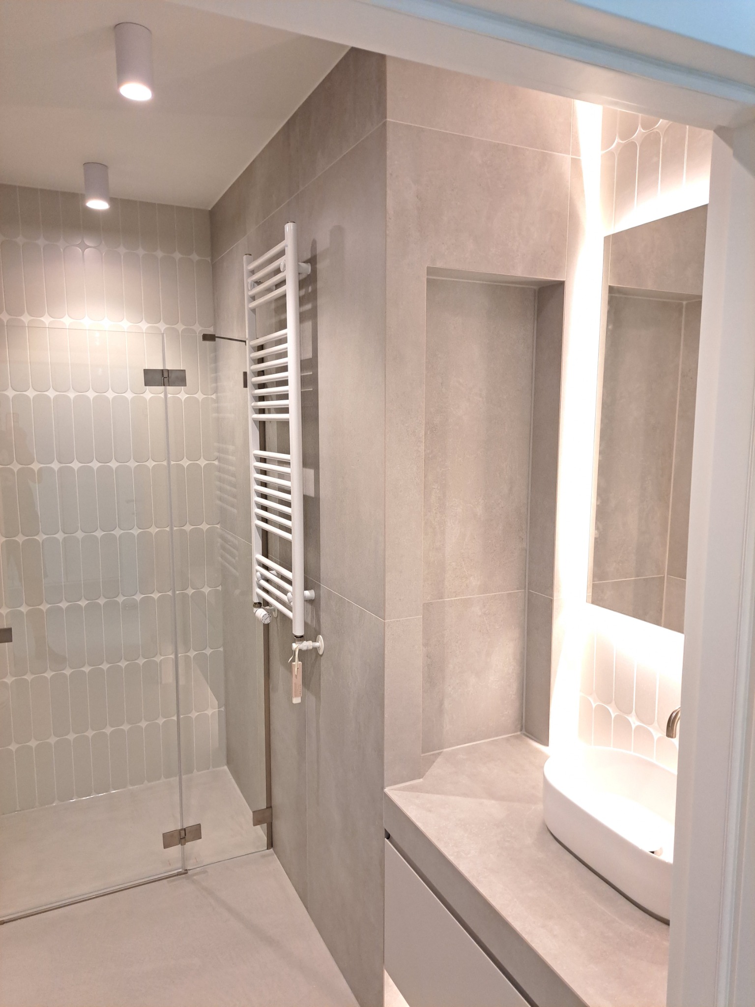 Nowoczesna łazienka z kabiną prysznicową ze szklanymi drzwiami, grzejnikiem drabinkowym i umywalką z podświetleniem LED za lustrem.