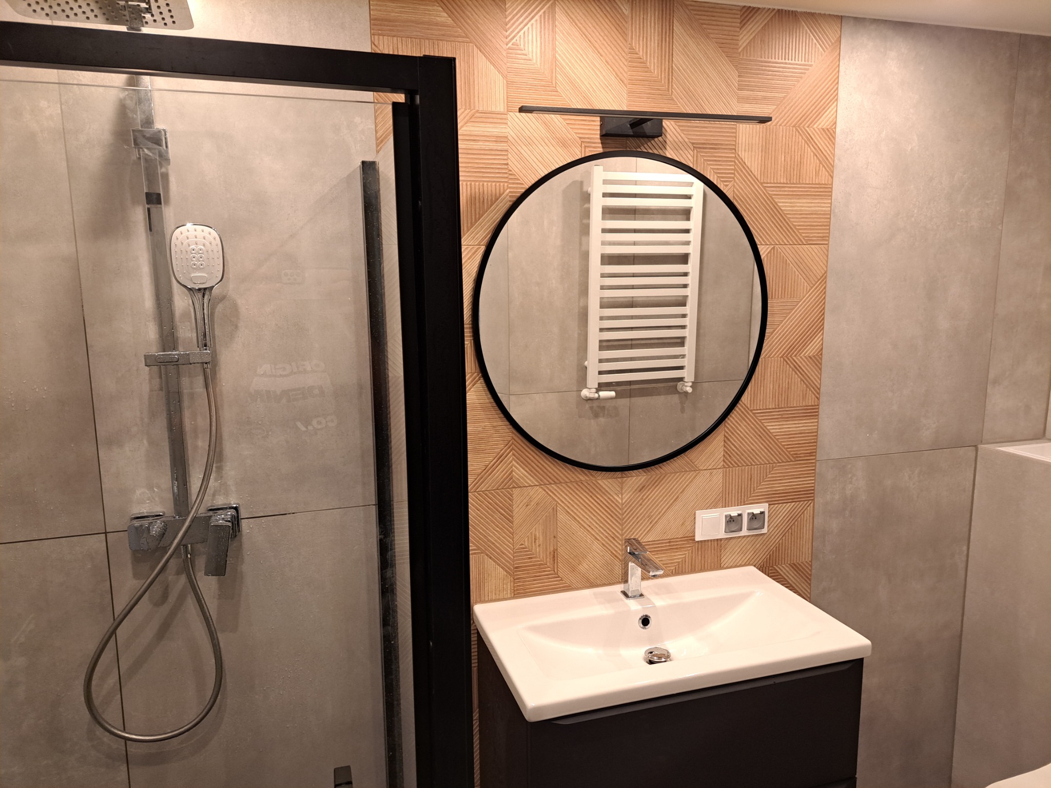 Nowoczesna łazienka we Wrocławiu z kabiną prysznicową ze srebrną armaturą, umywalką z czarną szafką, okrągłym lustrem z odbiciem grzejnika i dekoracyjną ścianą z drewnopodobnych płytek.