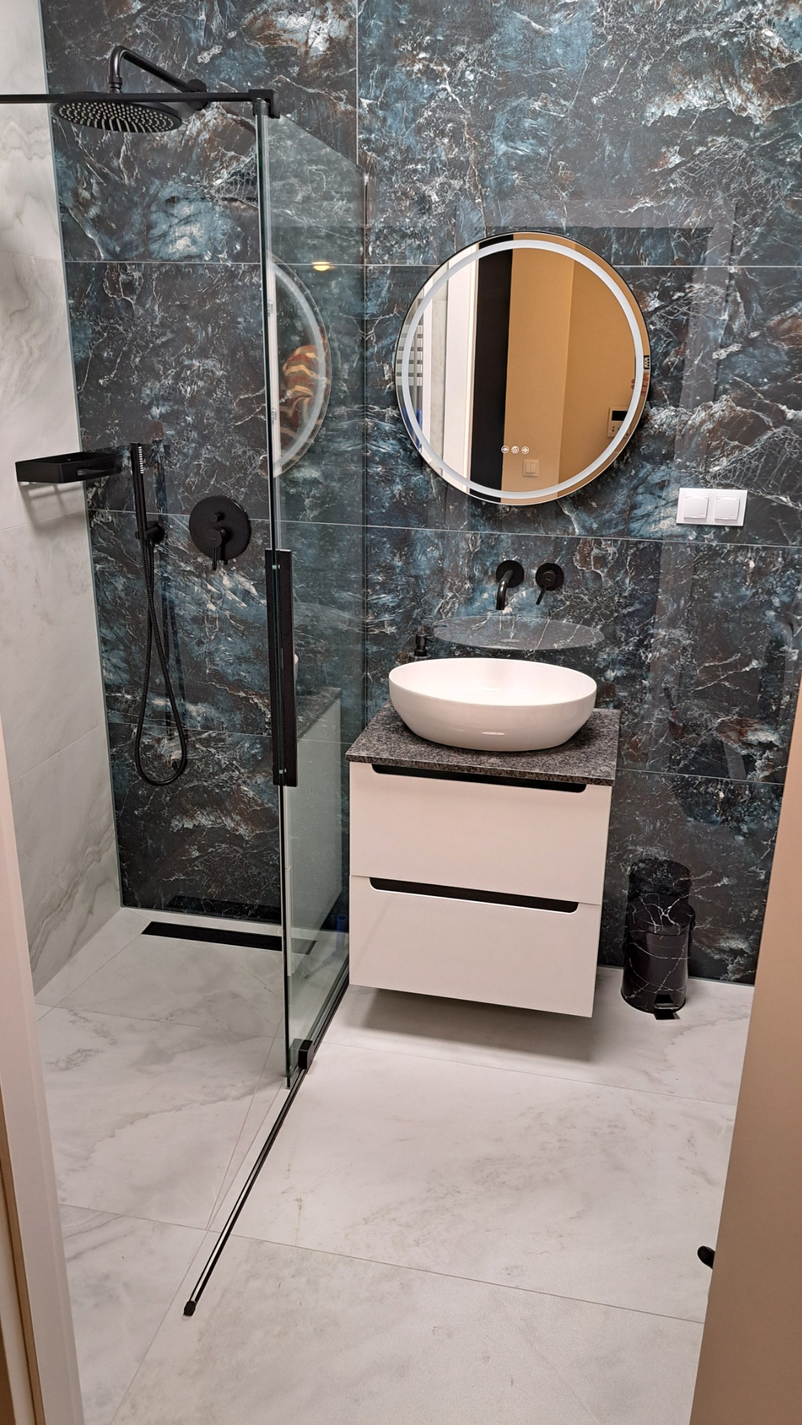 Nowoczesna łazienka z kabiną typu walk-in, umywalką na szafce z szufladami i okrągłym lustrem z podświetleniem. Płytki imitujące marmur w odcieniach granatu i bieli.
