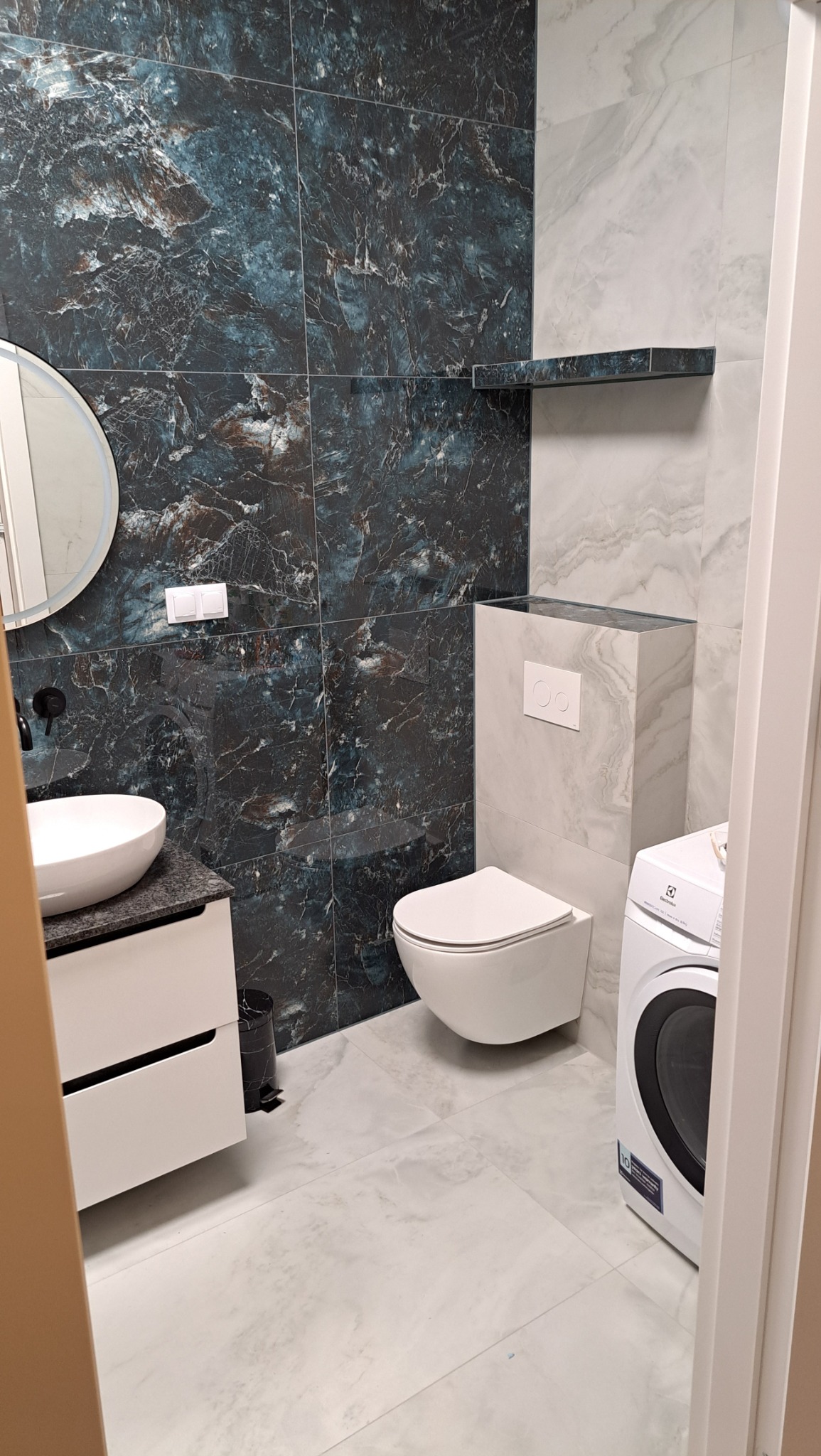 Elegancka łazienka z marmurowymi płytkami w odcieniach granatu i szarości. Umywalka nablatowa, toaleta podwieszana i pralka. Minimalistyczny design.