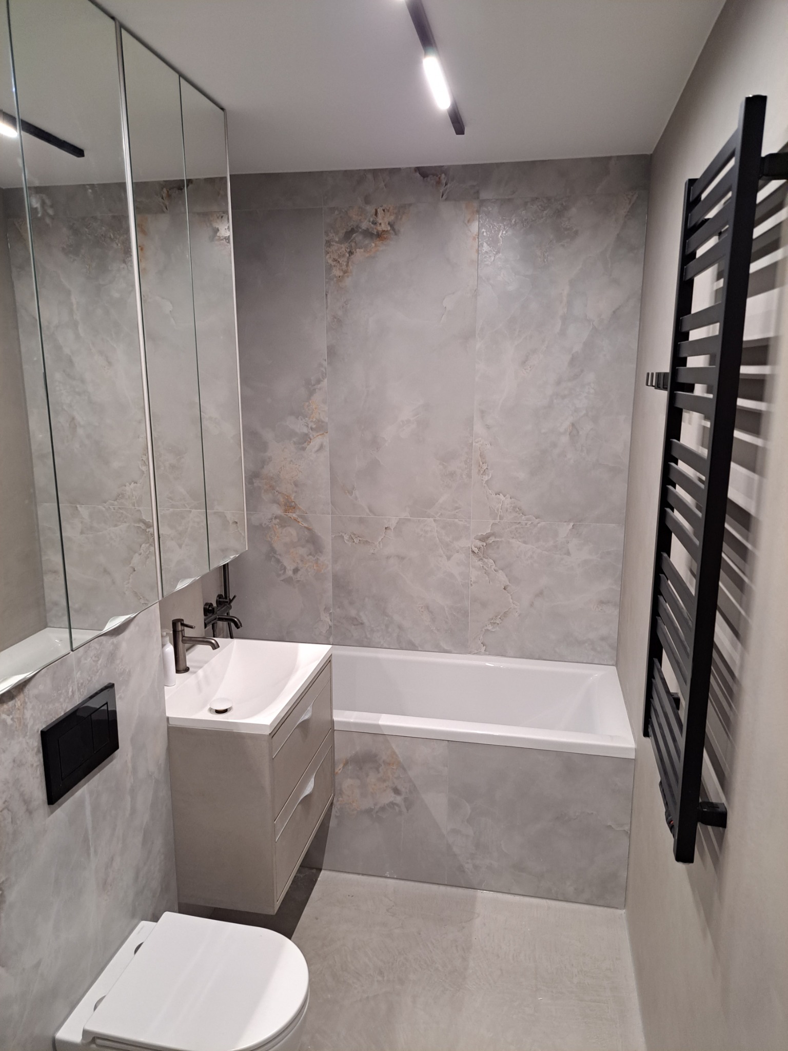 Nowoczesna łazienka z wanną obudowaną płytkami imitującymi marmur, wiszącą szafką z umywalką i czarnym grzejnikiem drabinkowym. Całość utrzymana w szarościach.