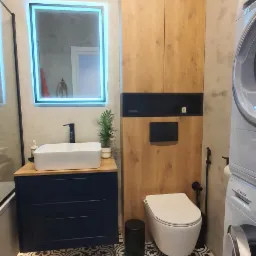 Nowoczesna łazienka z zabudową toalety, umywalką na szafce w kolorze granatowym, podświetlanym lustrem i wzorzystą podłogą.