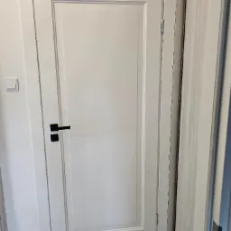 Białe, lakierowane drzwi wewnętrzne z czarną klamką, widoczne zawiasy i fragment sąsiedniej ściany z włącznikiem światła.