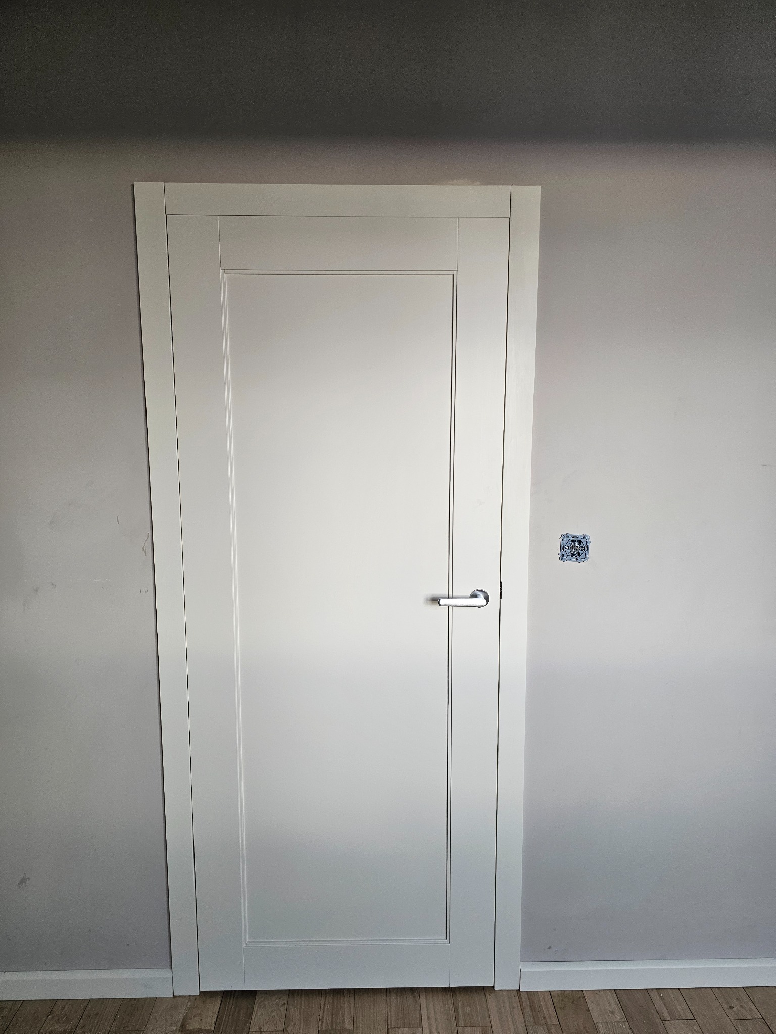 Białe drzwi wewnętrzne z prostokątną ramką i srebrną klamką, zamontowane w szarej ścianie z widoczną puszką elektryczną bez osprzętu.