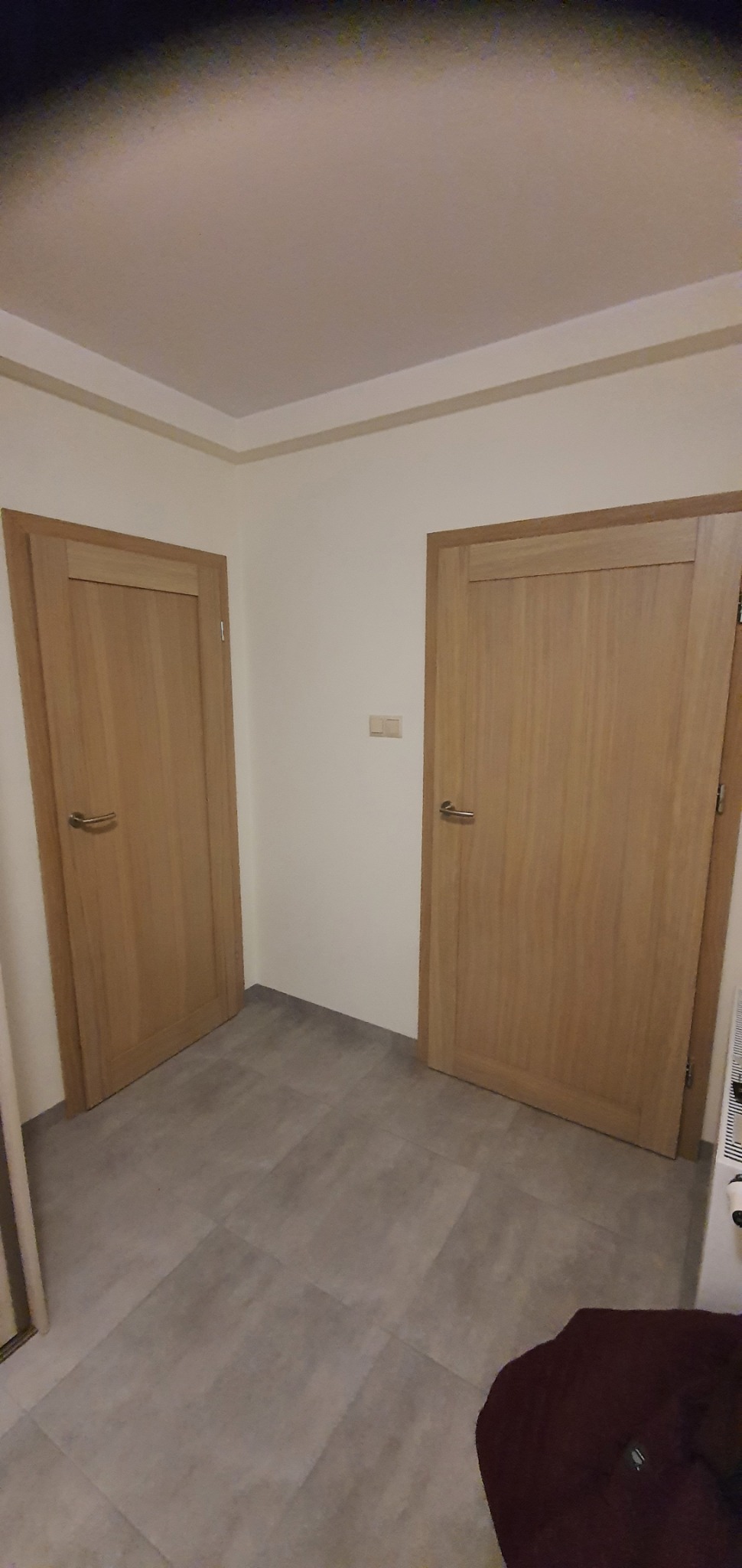 Dwa jasne, drewniane drzwi z metalowymi klamkami w białym pomieszczeniu z szarymi płytkami podłogowymi.