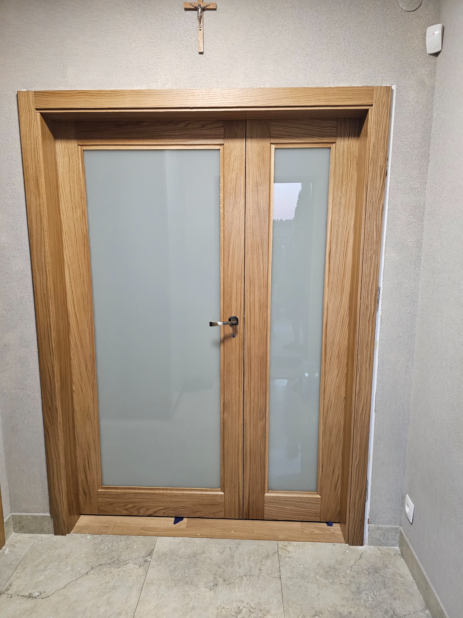 Dwuskrzydłowe drzwi z jasnego drewna z matowym szkłem, zamontowane w szarej ścianie, widoczny krzyż na ścianie powyżej.