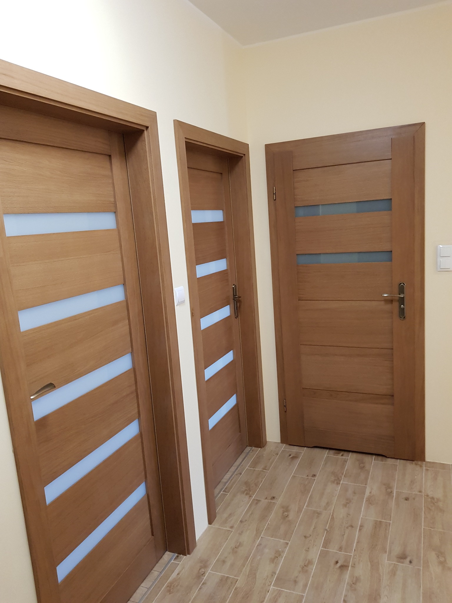 Trzy drewniane drzwi wewnętrzne z poziomymi przeszkleniami w odcieniach błękitu, zamontowane w jasnym korytarzu z podłogą imitującą drewno.