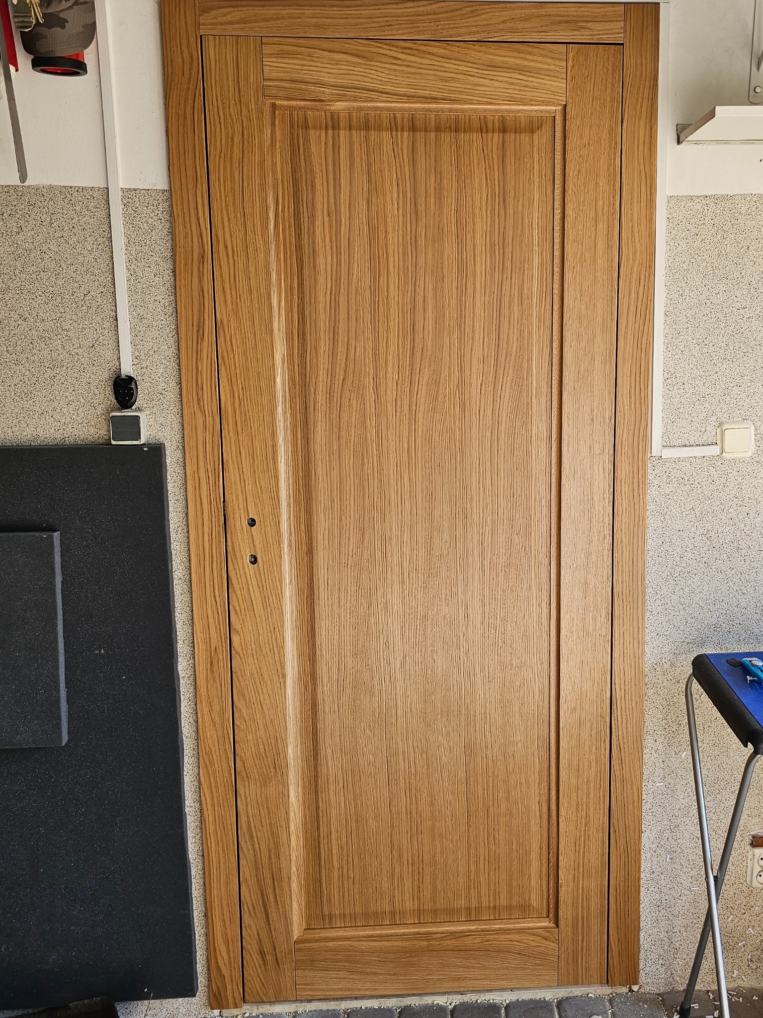 Zamknięte, drewniane drzwi wewnętrzne z widoczną strukturą drewna, osadzone w ościeżnicy, z dwoma małymi otworami na klamkę, na tle ściany o fakturze baranka.