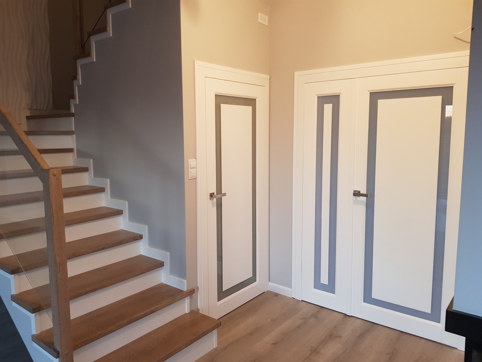 Dwie białe, nowoczesne drzwi drewniane z szarymi wstawkami, widoczne obok drewnianych schodów w jasnym wnętrzu.