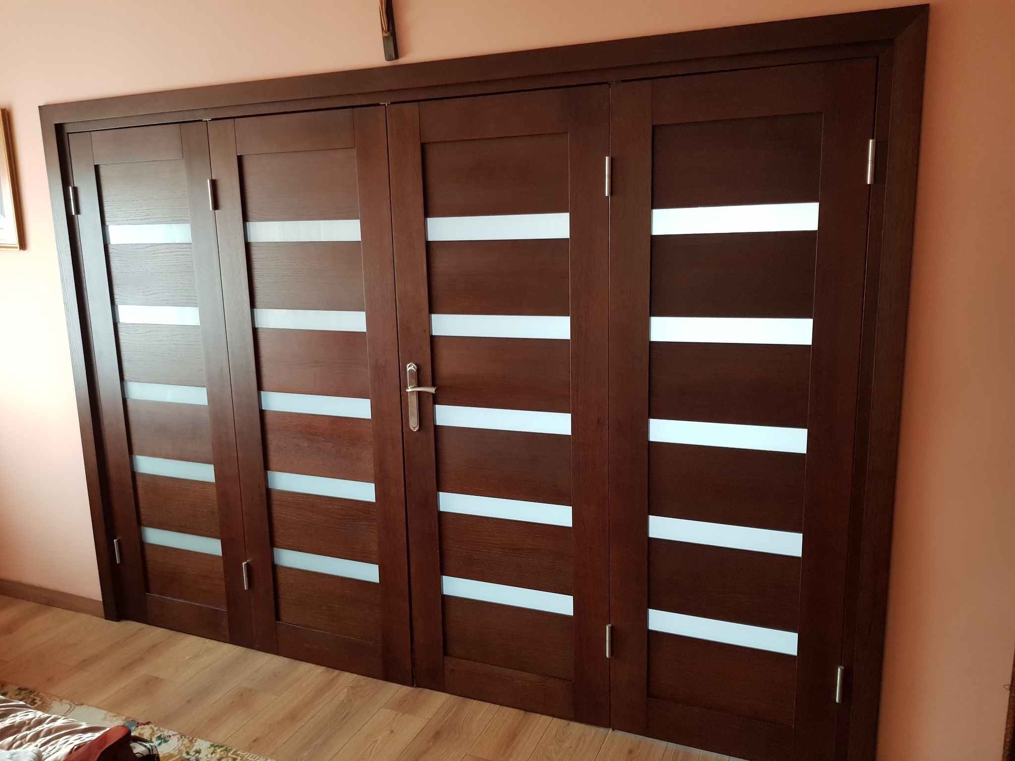 Czterodrzwiowe, ciemnobrązowe drzwi przesuwne z poziomymi, białymi wstawkami, widoczne na tle różowej ściany i drewnianej podłogi.