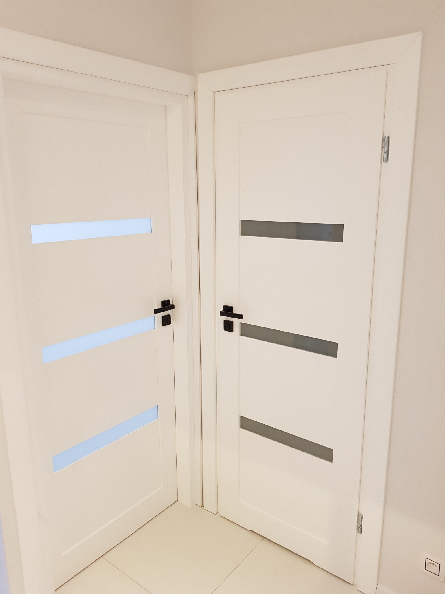 Dwa białe drzwi wewnętrzne z czarnymi klamkami i trzema poziomymi przeszkleniami, zamontowane w narożniku pomieszczenia z jasną podłogą.