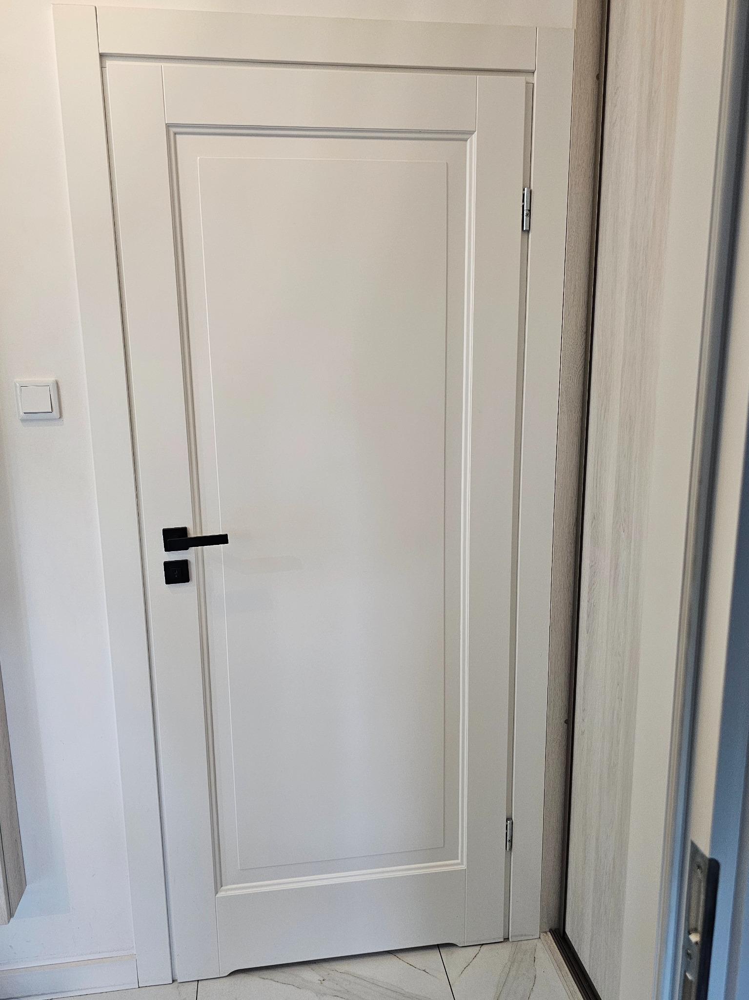 Białe, lakierowane drzwi wewnętrzne z czarną klamką, widoczne zawiasy i fragment sąsiedniej ściany z włącznikiem światła.