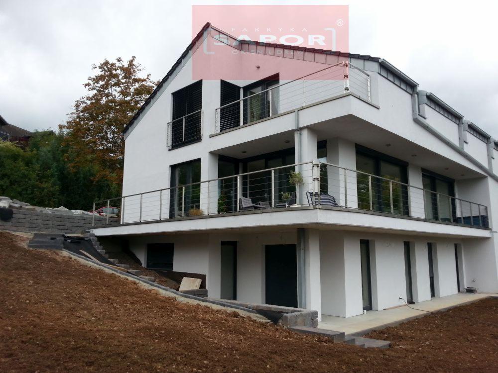 Nowoczesny, dwupiętrowy dom z białymi ścianami, balkonami z metalowymi balustradami i dużymi oknami, częściowo wykończony teren wokół budynku.