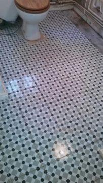 Wzorzysta podłoga w łazience z mozaiką w odcieniach szarości i czerni, widoczny fragment toalety z drewnianą deską i grzejnik żeberkowy w tle.