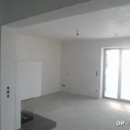 Wnętrze po nałożeniu tynku maszynowego, widoczne gładkie ściany i sufit, przygotowane do dalszych prac wykończeniowych, z widocznymi puszkami elektrycznymi i oknem w tle.