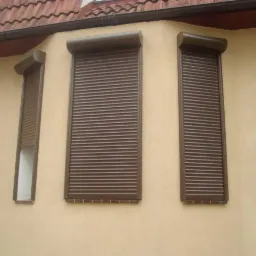 Brązowe rolety zewnętrzne zamontowane na elewacji domu z beżowym tynkiem i czerwoną dachówką, jedna roleta częściowo podniesiona.