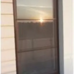 Brązowe okno z zamontowaną moskitierą, widok z zewnątrz, zachód słońca odbijający się w szybie.