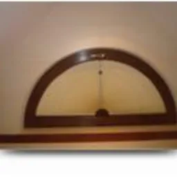 Półokrągłe okno z brązową ramą zamontowane w jasnej ścianie, widoczne delikatne refleksy światła.