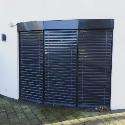 Zewnętrzne żaluzje fasadowe w kolorze antracytowym, zamontowane na białej fasadzie budynku, widoczne z poziomu chodnika z kostki brukowej.